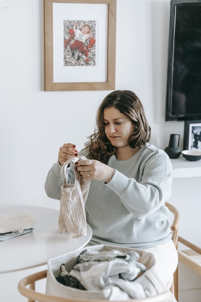 Concentrated female with baby garment in hands sitting at white table near bag of clothes in light room at home